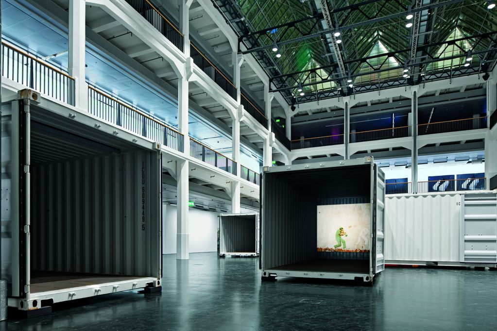 Installationsansicht, die mindestens drei weiße offene Container in verschiedenen Positionen in einer großen Halle zeigt. In einem davon ist eine Videoprojektion des Künstlers Robin Rhode zu sehen. Das Standbild stellt einen Mann in einem hellgrünen Outfit auf dem Boden dar, auf den ein Backsteinziegel zufliegt. Robin Rhode, Sammlung Goetz München.