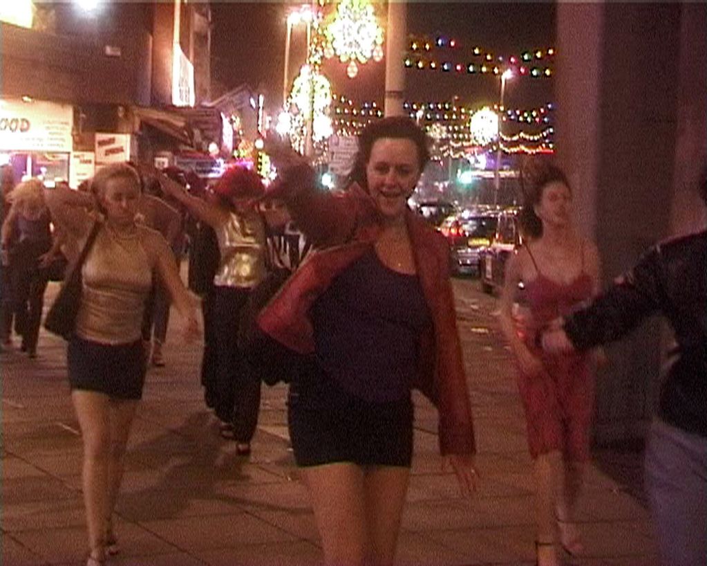 Group of young women in a party mood on the street. They wear short skirts, partly shimmering clothes and wigs. In the foreground, one of them stretches out her arm toward the camera/viewer cheerfully.