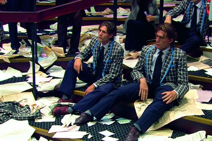 Diese Aufnahme zeigt ein Filmstill von drei gleich gekleideten Männern, mit den gleichen Frisuren, die erschöpft und resigniert auf breiten Stufen sitzen. Der Boden ist voll mit verstreuten Papieren, deren Inhalt nicht erkennbar ist. Aernout Mik,Sammlung Goetz München