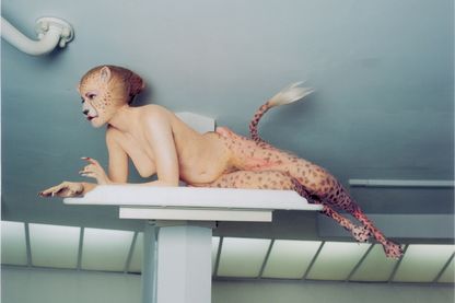 Diese Aufnahme zeigt eine halb liegende Leoparden-Mensch-Dame, die die Beine, das Gesicht und den Schwanz eines Leoparden aufweist und sonst menschliche Attribute besitzt. An ihrem Maul und ihren falschen, krallenartigen Fingernägeln ist Blut zu erkennen. Sie befindet sich in einem hell ausgeleuchteten weißen Raum auf einem Sockel.