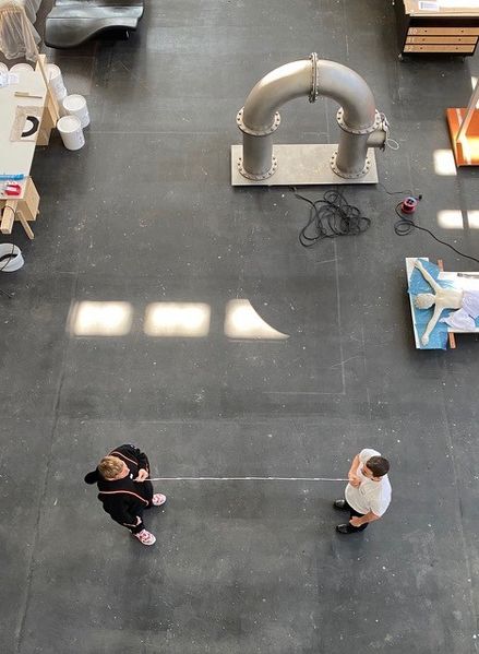 Vogelperspektive auf die zwei Künstler in ihrem Atelier, wie sie den Abstand zwischen ihnen mit einer Schnur messen