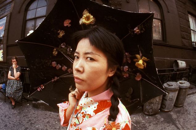 Eine junge asiatische Frau in einem Kimono. Sie trägt einen Regenschirm, auf dessen innenseite Plastikblumen kleben.