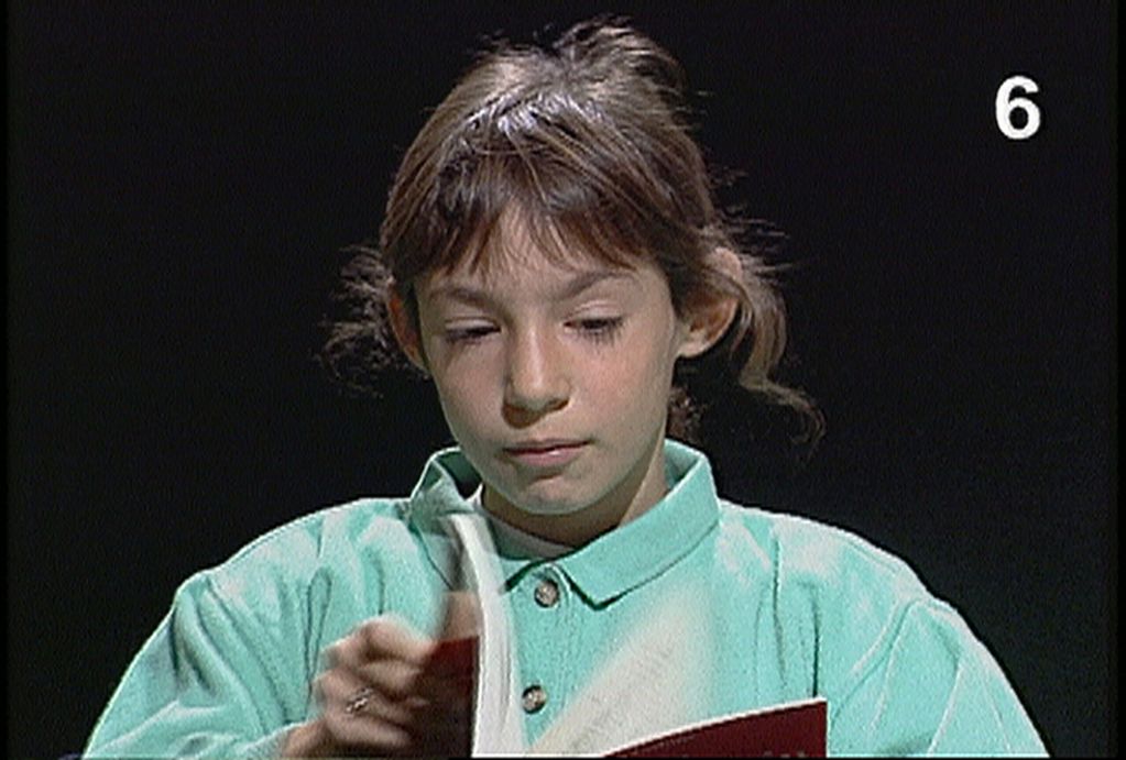 Auf diesem Videostill sieht man ein etwa zehnjähriges Mädchen mit hellbraunen Haaren, das aus einer roten Ausgabe des Buches "Bemerkungen über die Farben" von Ludwig Wittgenstein vorliest. 