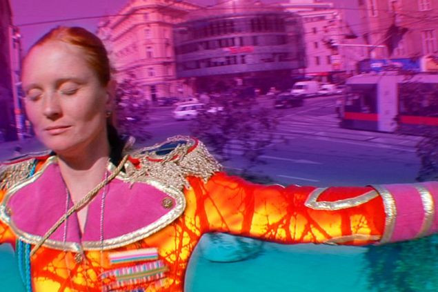 Eine Frau mit ausgebreiten Armen vor pinkfarbenem Hintergrund. Sie trägt ein buntes orangenfarbenes Kostüm, das mit dem Hintergrund kontrastiert und hat geschlossene Augen.