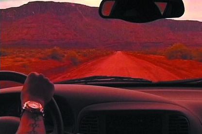 Video Still einer in rosarotem Licht getauchten Autofahrtszenerie, von der Rückbank durch die Frontwindschutzscheibe aus gesehen. Olaf Breuning, Sammlung Goetz München