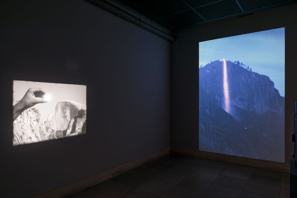 Installation view with two projections. Left: a hand holds an object reflecting the sunlight against the background of a mountain range. On the right: Nightly shot of a mountain wall. A bright light steel shines down from the mountain into the valley. Cyrill Lachauer, Sammlung Goetz, Munich