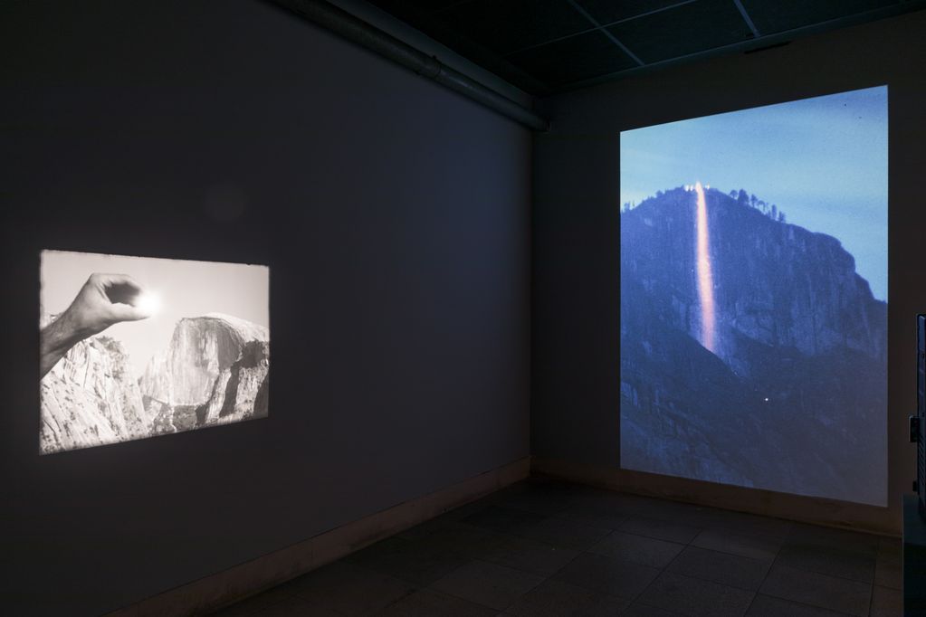 Installatiionsaufnahme mit zwei Projektionen. Links: eine Hand hält ein das Sonnenlicht reflekierendes Objekt vor dem Hintergrund einer Bergkette. Rechts: Nächtliche Aufnahme einer Bergwand. Ein heller Lichtstahl scheint vom Berg ins Tal herunter. Cyrill Lachauer, Sammlung Goetz, München