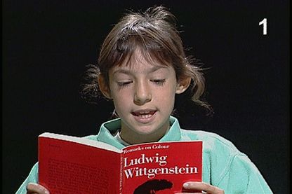 Auf diesem Videostill sieht man ein etwa zehnjähriges Mädchen mit hellbraunen Haaren, das aus einer roten Ausgabe des Buches "Bemerkungen über die Farben" von Ludwig Wittgenstein liest. 