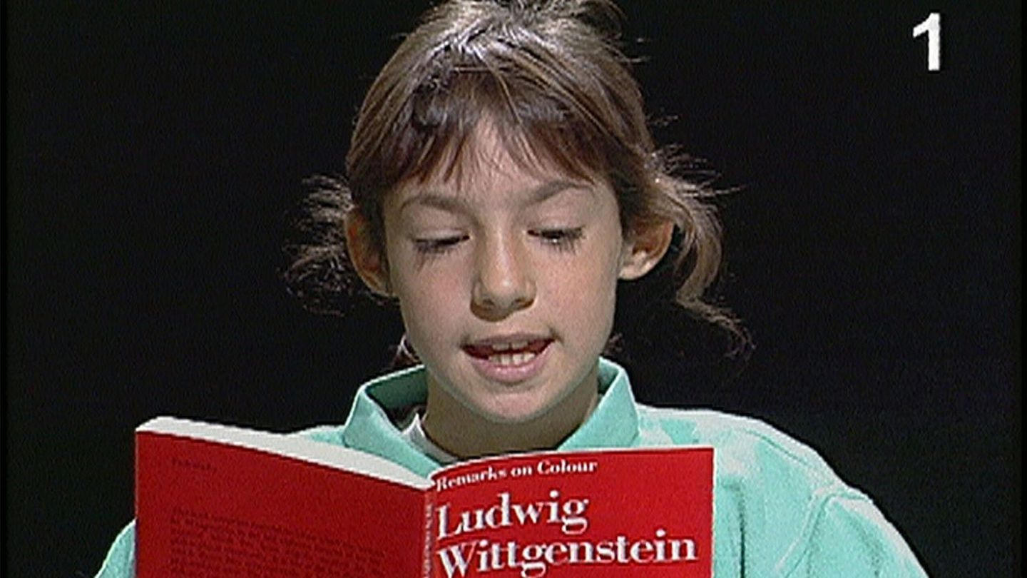 Auf diesem Videostill sieht man ein etwa zehnjähriges Mädchen mit hellbraunen Haaren, das aus einer roten Ausgabe des Buches "Bemerkungen über die Farben" von Ludwig Wittgenstein liest. 