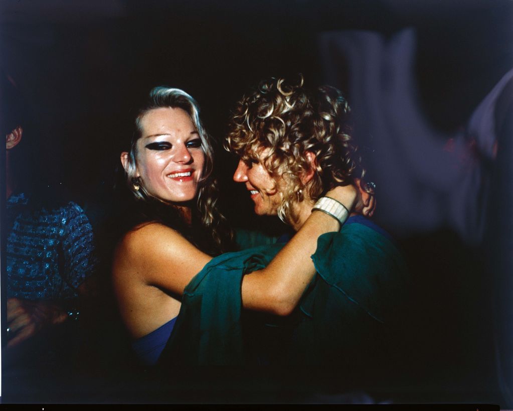 In this colour photograph two women hold each other in their arms in a club. The right woman with blonde curly hair can be seen in half profile, while the other blonde and heavily made-up woman looks laughing into the camera.