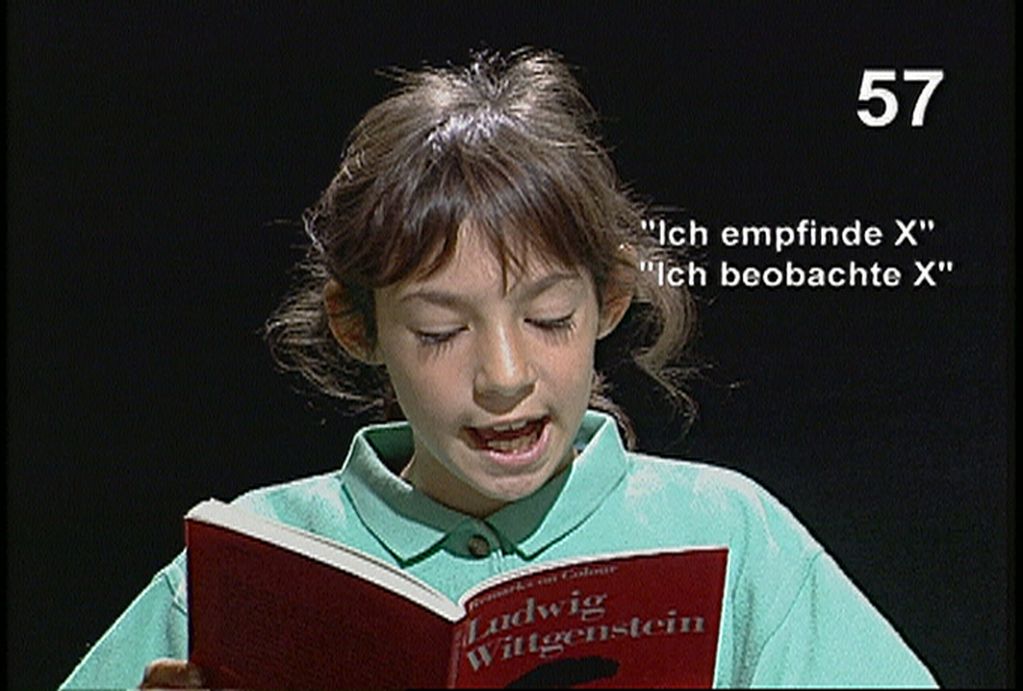 Auf diesem Videostill sieht man ein etwa zehnjähriges Mädchen mit hellbraunen Haaren, das aus einer roten Ausgabe des Buches "Bemerkungen über die Farben" von Ludwig Wittgenstein vorliest. 