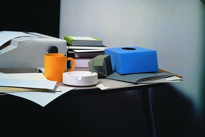 In this colour photograph, an office table with objects on it can be seen. The complete ensemble is made of paper. The objects are a typewriter, a black, narrow folder with documents on it, a stack of books in different colours and sizes, an orange-yellow cup with a spoon in it, paper, notebooks, an empty box of tissues and a small box of paper clips.