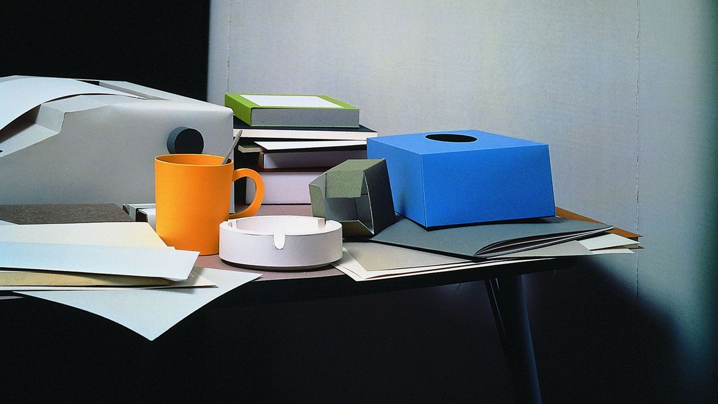 In this colour photograph, an office table with objects on it can be seen. The complete ensemble is made of paper. The objects are a typewriter, a black, narrow folder with documents on it, a stack of books in different colours and sizes, an orange-yellow cup with a spoon in it, paper, notebooks, an empty box of tissues and a small box of paper clips.