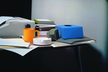 Auf dieser Farbfotografie ist ein Bürotisch mit Objekten darauf zu sehen. Das komplette Ensemble besteht aus Papier. Bei den Objekten handelt es sich um eine Schreibmaschine, einem schwarzen, schmalen Ordner mit Dokumenten darauf, einem Stapel Bücher in verschiedenen Farben und Größen, einer orange-gelben Tasse mit Löffel darin, Papier, Hefte, eine leere Taschentuchbox und eine kleine Büroklammerschachtel.