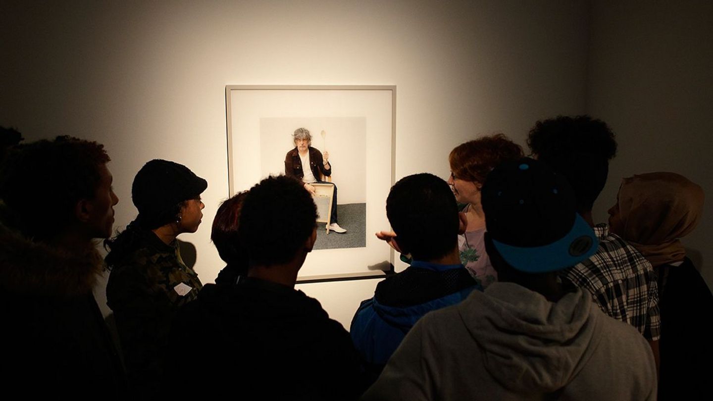 Eine Gruppe junger Menschen steht im Halbreis um einen Wandabschnitt an dem eine Fotografie hängt