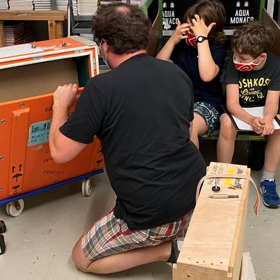 Ein Mann zeigt zwei Kindern eine Kunst-Transportbox. Link zum Workshop Discover it