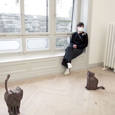 © Zilla Leutenegger Fotografie der Künstlerin mit Mund-Nasen-Schutz. Vor Ihr auf dem Boden sind zwei Katzenskulpturen aus Bronze. Sammlung Goetz, München