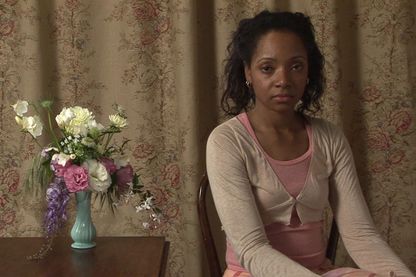 Video Still einer jungen schwarzen Frau, die vor einem geblümten Stoff sitzt, neben ihr auf dem Holztisch steht eine zierliche, hellblaue Vase mit pastellfarbenen Blumen darin. Sie selbst sieht resigniert in die Kamera. Andrea Bowers, Sammlung Goetz München