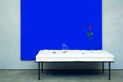 This installation shot seems like a three-dimensional still life. A blue, monochrome painted canvas serves as a background. In the foreground is a metal bedstead with a white, uncovered mattress on it. On the mattress are, from left to right: two nuts, a carafe of water with a fish in it and an upright rose.