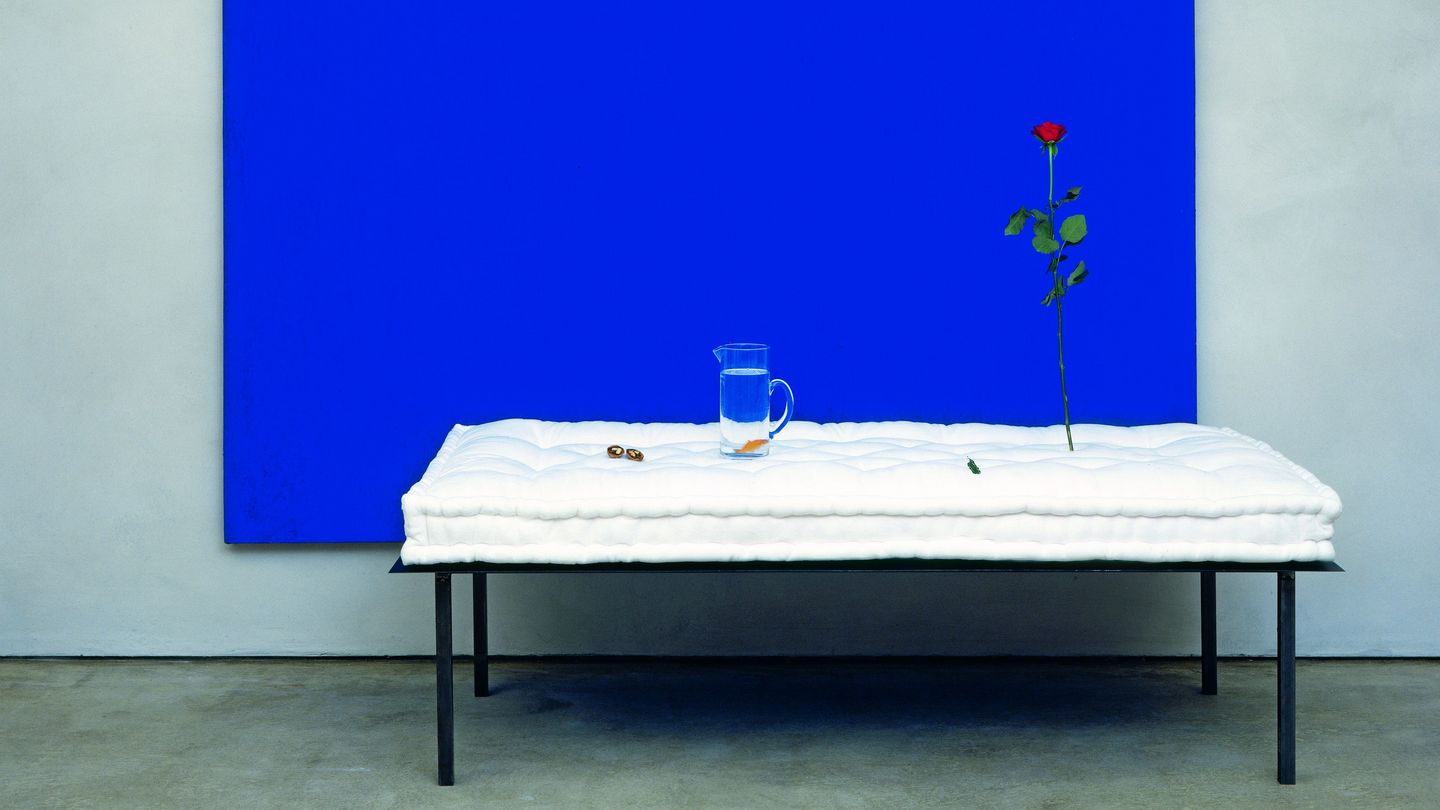 This installation shot seems like a three-dimensional still life. A blue, monochrome painted canvas serves as a background. In the foreground is a metal bedstead with a white, uncovered mattress on it. On the mattress are, from left to right: two nuts, a carafe of water with a fish in it and an upright rose.