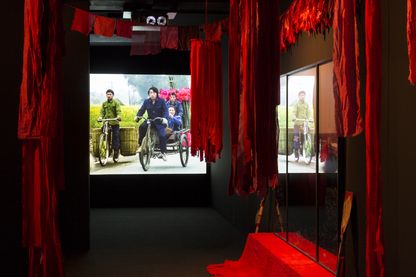 Detail der Installation der Arbeit "Floating Food"; zu sehen sind von der Decke hängende rote Stoffbahnen in einem dunklen Raum sowie ein Video Still, das junge asiatische Männer in traditioneller Arbeitskleidung auf Fahrrädern auf einer Landstraße zwischen grünen Feldern zeigt. Ulrike Ottinger, Sammlung Goetz München 