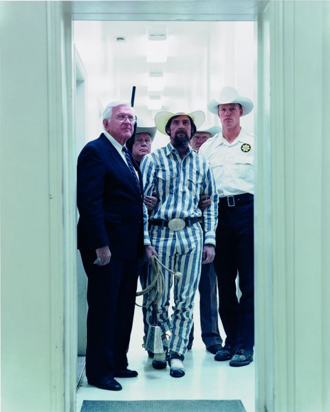 This film still shows men in suits with cowboy hats in a corridor. The man in the middle wears a grey and white striped prisoners suit, holding a rope and a cowbell in his hand, but is grabbed by two other men by the arms.