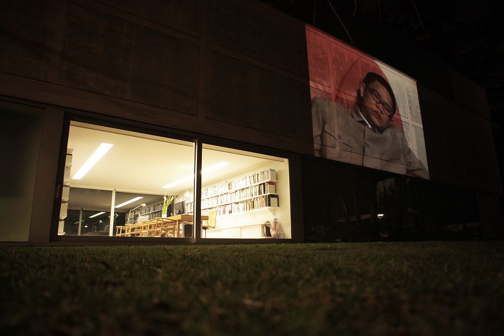Installationsansicht der Videoarbeit "Honey mi" des Künstlers Yang Fudong auf der Fassade des Gebäudes der Sammlung Goetz. Zu sehen ist der hell erleuchtete Eingangsbereich sowie das Standbild eines asiatisch aussehenden Mannes mittleren Alters mit geschlossenen Augen vor rot-weißem Hintergrund. Yang Fudong, Sammlung Goetz München