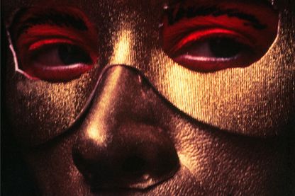 Diese Fotografie der Künstlerin Cindy Sherman zeigt eine Nahaufnahme ihres golden geschminktem Gesichts, welches mit einer goldfarbenen Augenmaske teilweise bedeckt wird. Die Augen sind hellrot geschminkt, die Augenbrauen sind schwarz nachgemalt, sie blinkt nach rechts und hat einen schwarzen Strich auf den Lippen, der über die Länge ihrer eigentlichen Lippen hinausgeht.