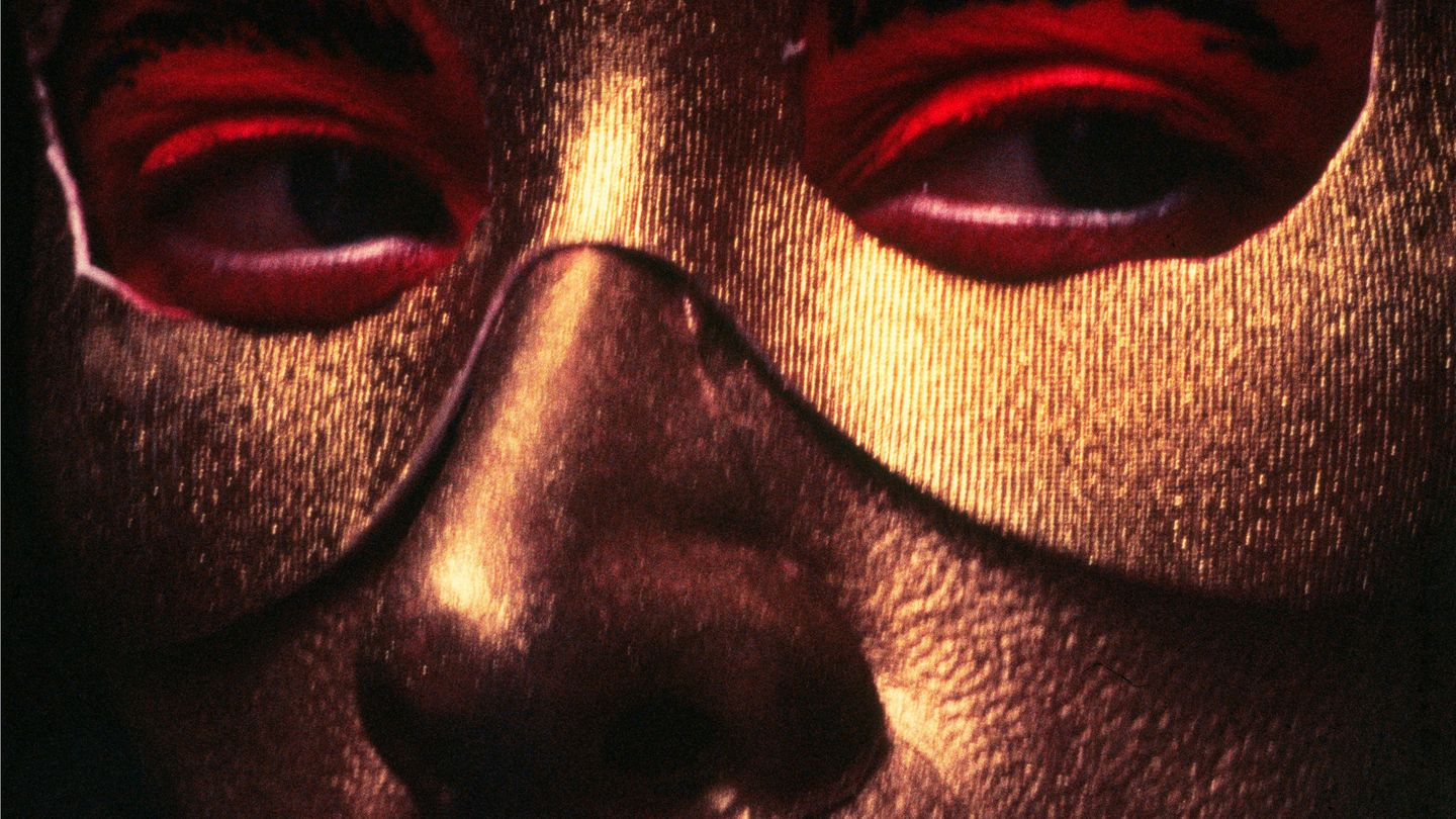 This photograph by the artist Cindy Sherman shows a close-up of her golden painted face, which is partially covered with a golden eye mask. The eyes are painted light red, the eyebrows are painted black, she is blinking to the right and has a black line on her lips that extends beyond the length of her actual lips.